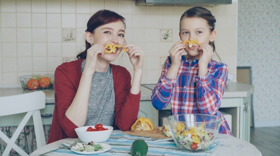 Joyful mother and cute cheerful daughter have fun grimacing silly with vegetables and looking into camera while cooking in the kitchen at home. Family, cook, and people concept